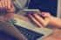 A person sitting at a desk in front of a laptop while holding a smartphone in their left hand.