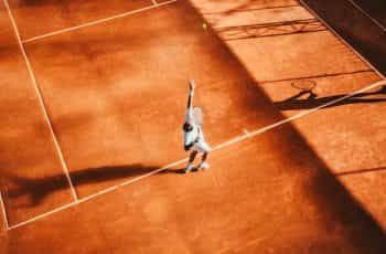 A man in a white tennis outfit serves a ball standing on an orange clay court.